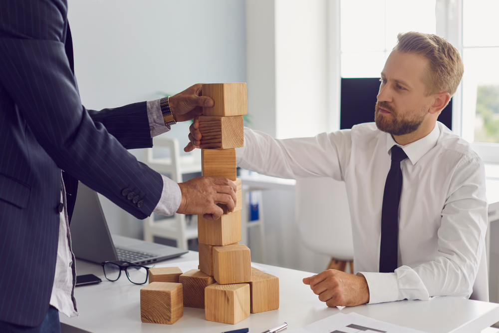 Two business professionals collaboratively building a tower of wooden blocks in an office, symbolizing the concept of resilience and teamwork in the workplace.