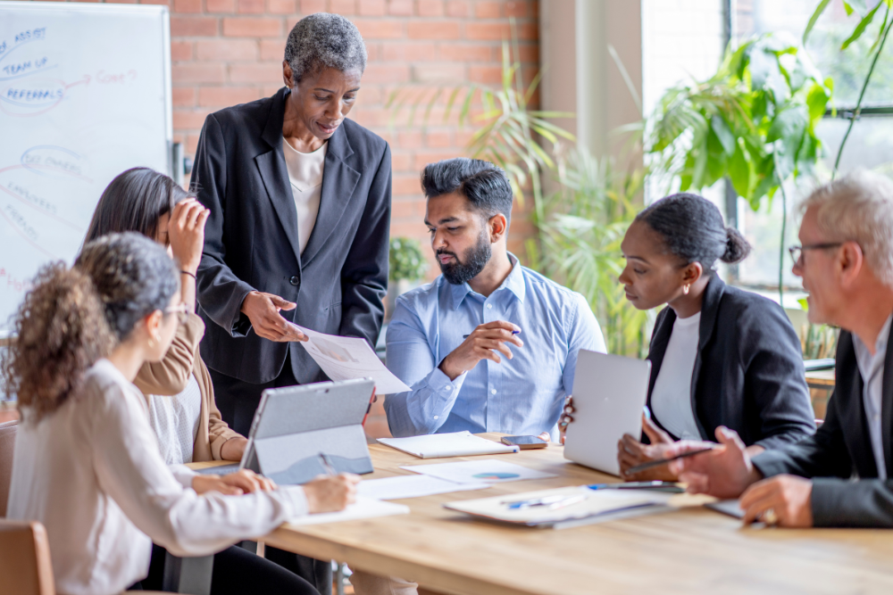 A diverse group of professionals collaborating during a meeting, with a senior leader providing guidance, symbolizing teamwork, leadership, and learning in the workplace.